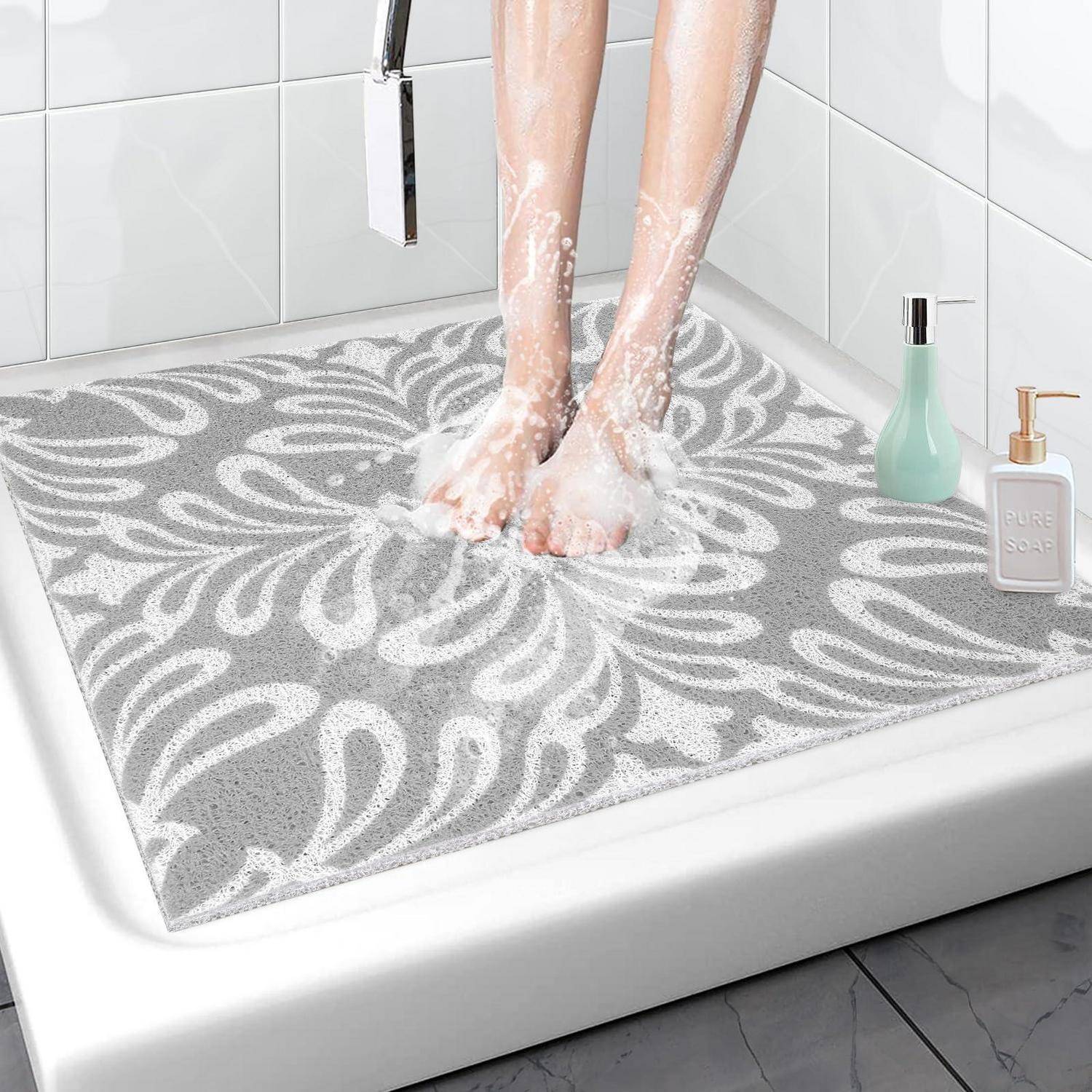 Person wäscht sich die Füße auf einer grauen floralen Badmatte in einer Dusche, mit Seifenflaschen daneben.