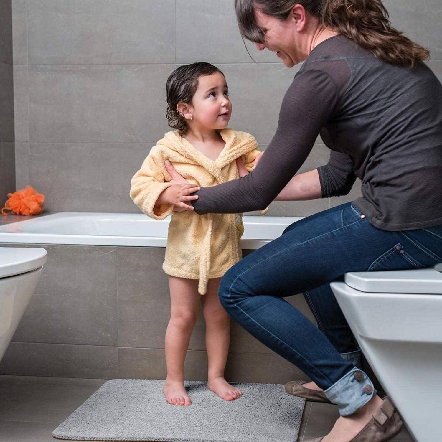 Eine Frau hilft einem kleinen Kind im Badezimmer neben einer Badewanne zu stehen und hält dabei die Arme des Kindes zur Unterstützung.