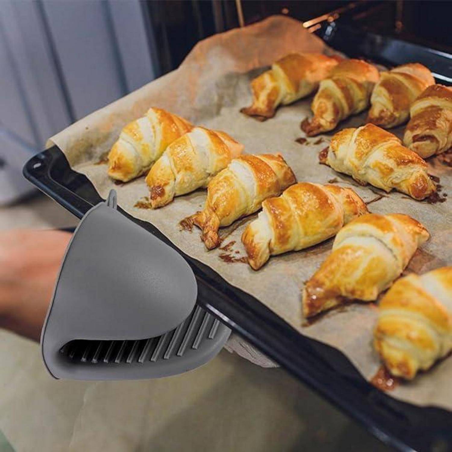 Tablett mit frisch gebackenen Croissants auf Pergamentpapier, wobei ein graues Silikon-Ofenhandschuh die heiße Backform hält.