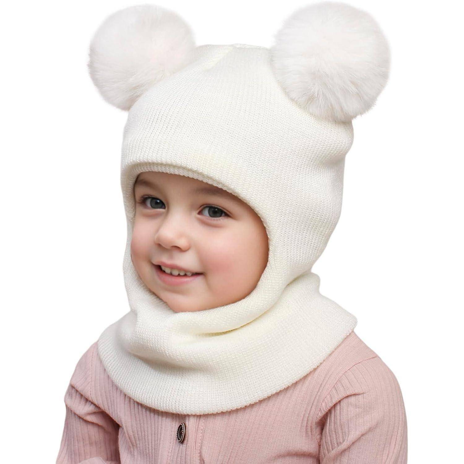 Lachendes Kind, das eine weiße gestrickte Wintermütze mit zwei Pompons und einen passenden Schal trägt, vor einem einfachen weißen Hintergrund.