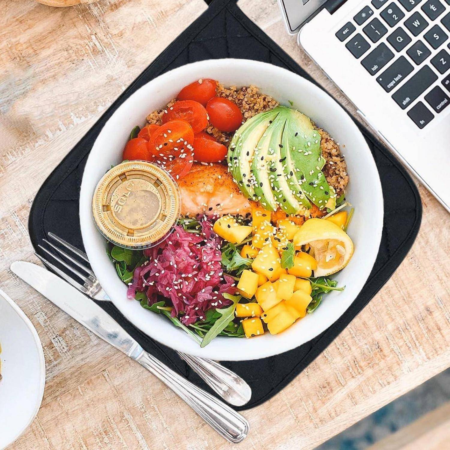 Draufsicht auf eine farbenfrohe Poke-Schüssel mit Avocado, Lachs, Tomaten, Mango, Quinoa, Grünzeug und einer Saucenkanne auf einer Serviette neben einem Laptop und Besteck.