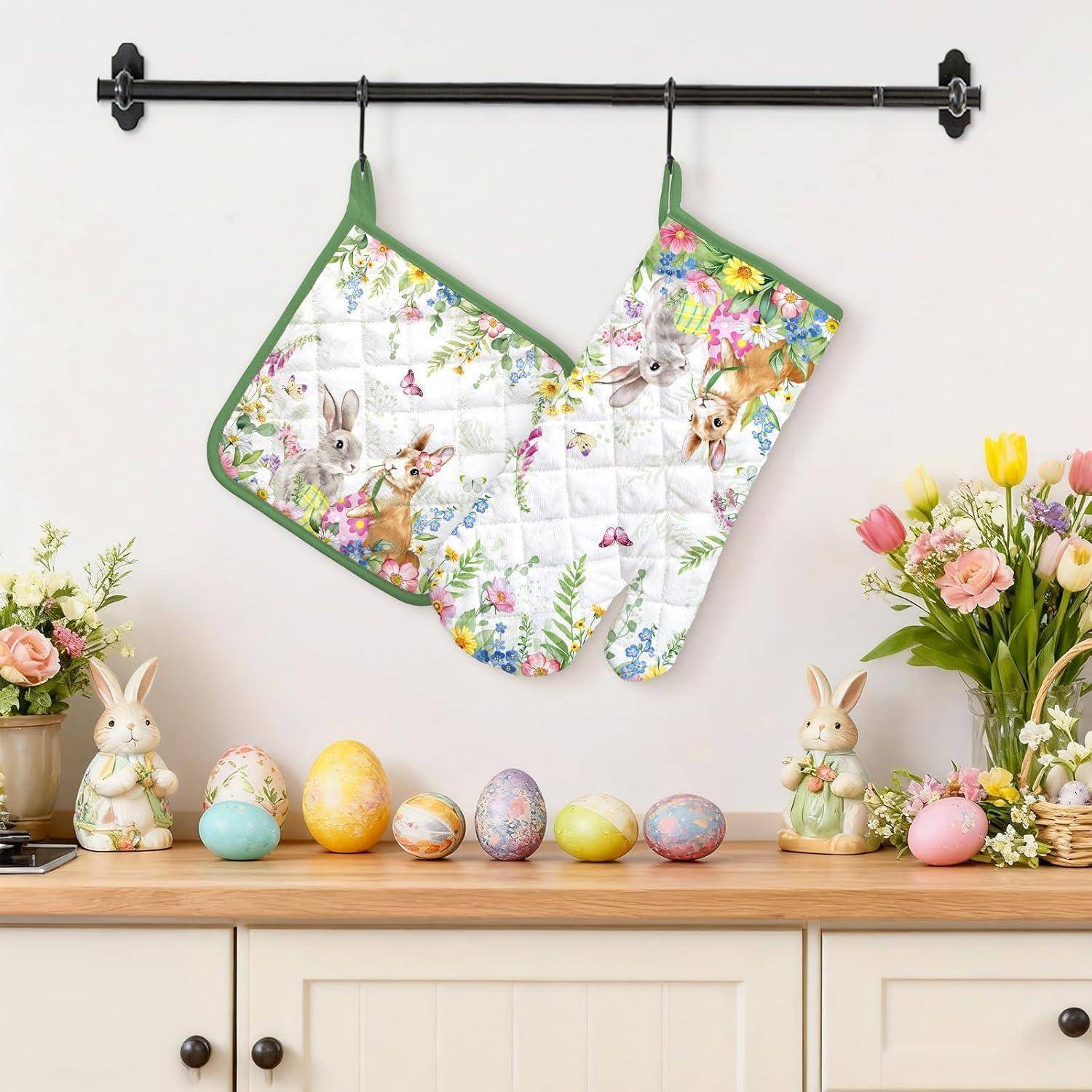 Zwei blütengesteppte Ofenhandschuhe hängen an einer Wand über einer Kommode mit pastellfarbenen Ostereiern, Hasenfigruen und Blumen.