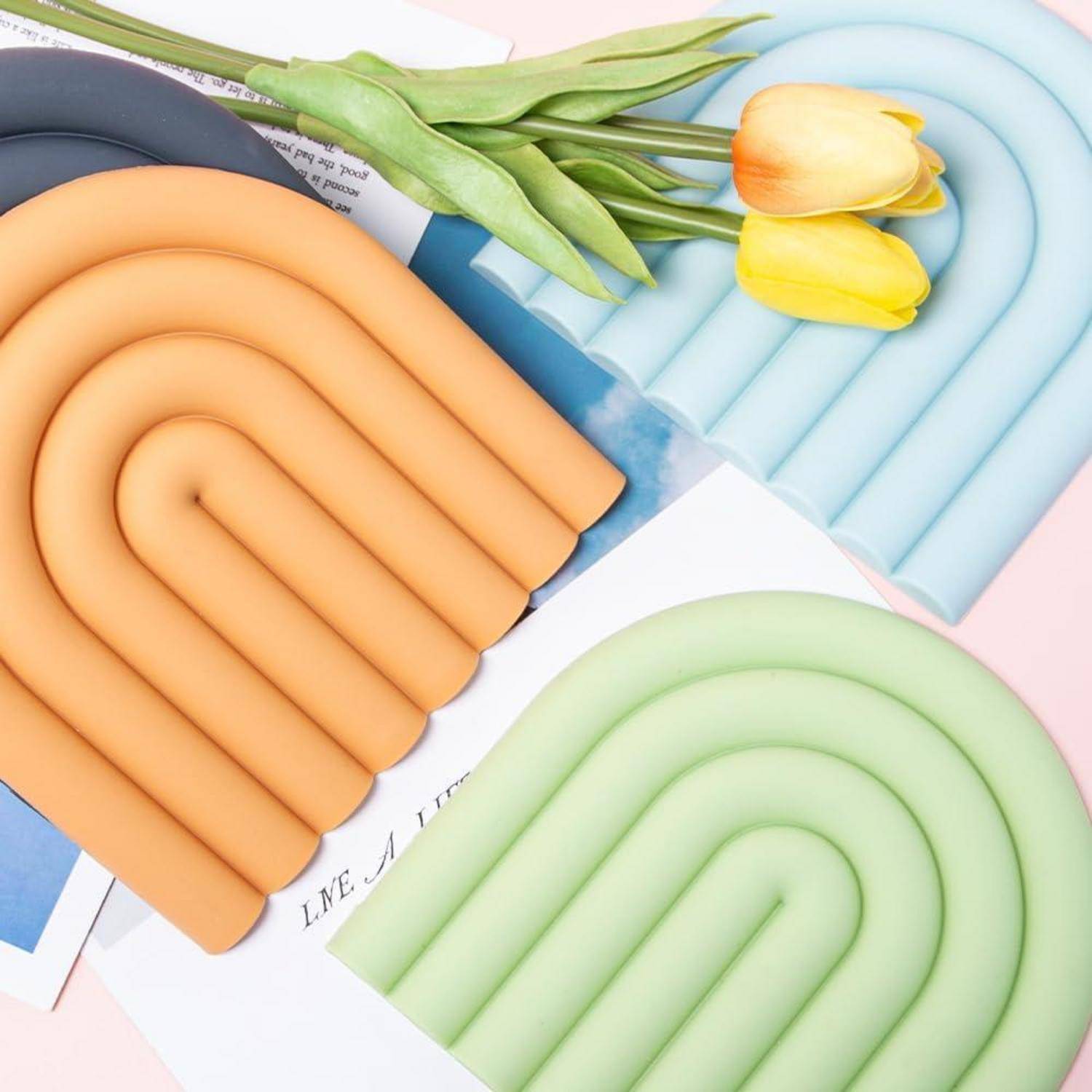 Vier pastellfarbene regenbogenförmige Silikonuntersetzer in Orange, Blau, Grün und Gelb auf Zeitschriften mit Tulpen angeordnet.