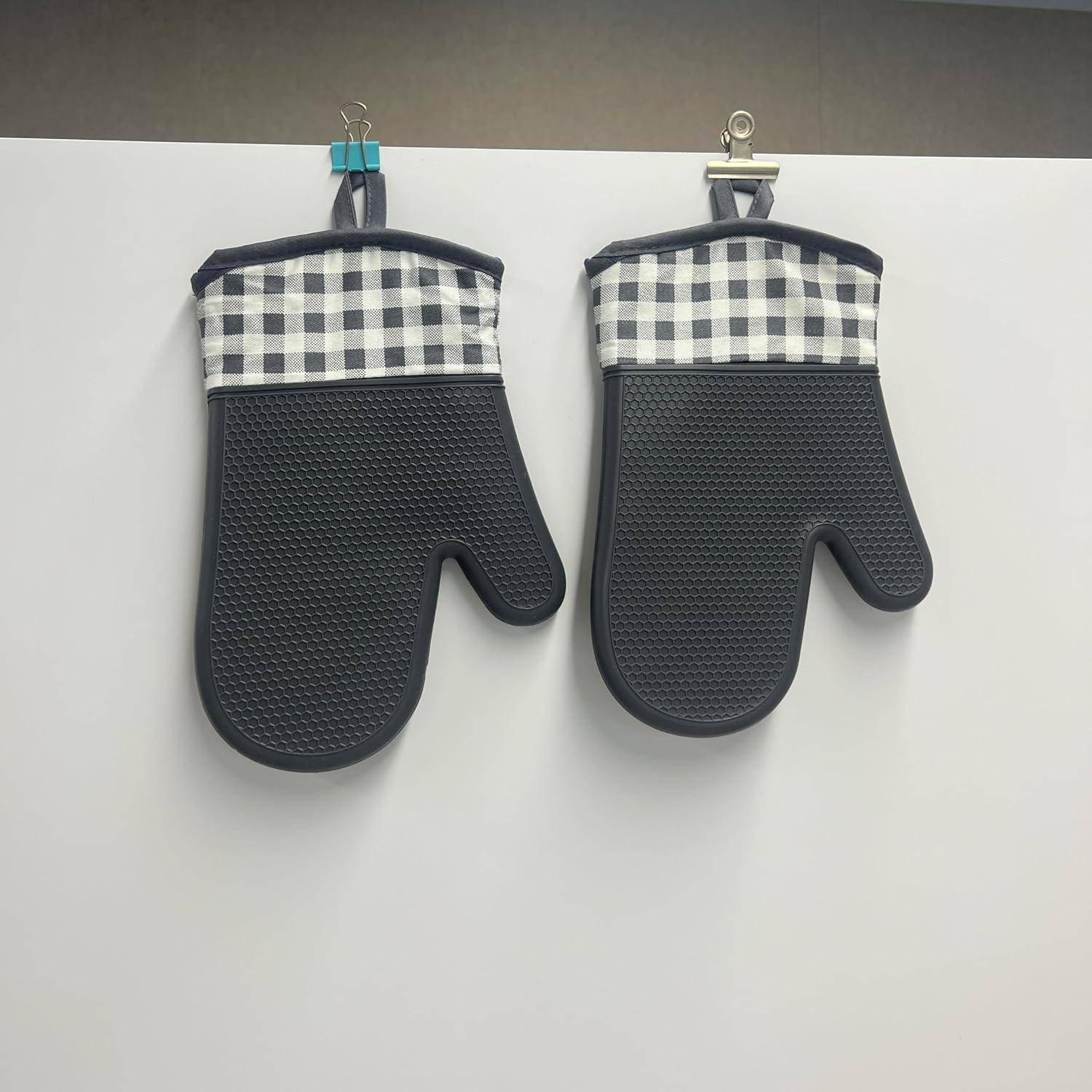Zwei schwarze Ofenhandschuhe mit schwarz-weißen karierten Manschetten hängen nebeneinander auf Wandclips.