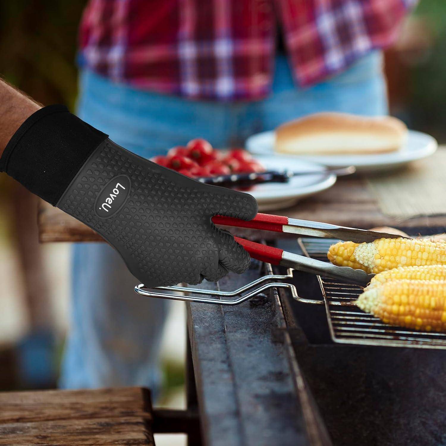 Behandschuhte Hand, die rote Zangen benutzt, um Mais auf einem Grill zu wenden, mit einer anderen Person und verschwommenem Picknick-Essen im Hintergrund.