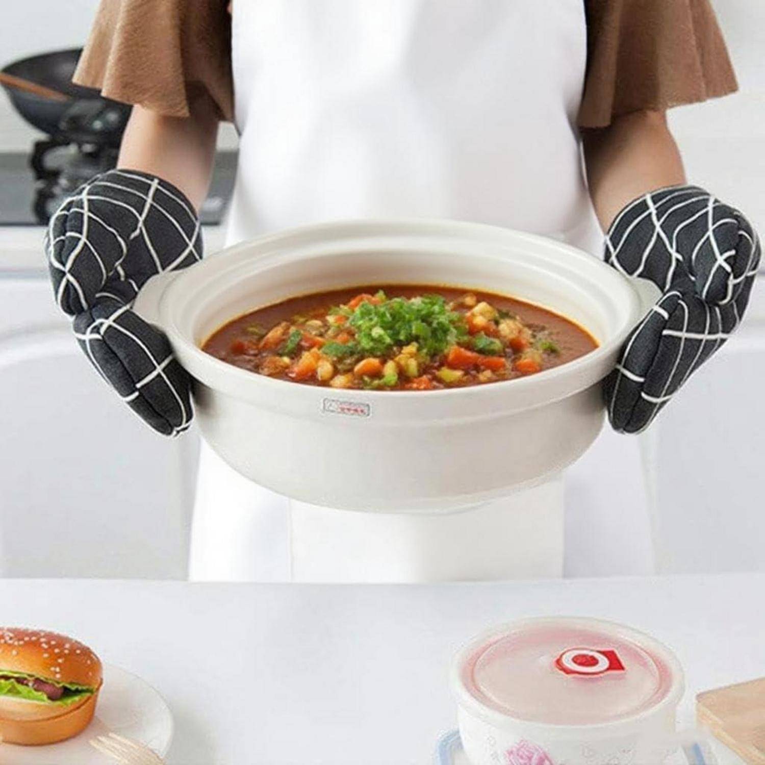Person in Küchenhandschuhen, die eine weiße Suppenschüssel mit Gemüsesuppe hält, mit einem Behälter mit Deckel und einem Burger im Hintergrund.