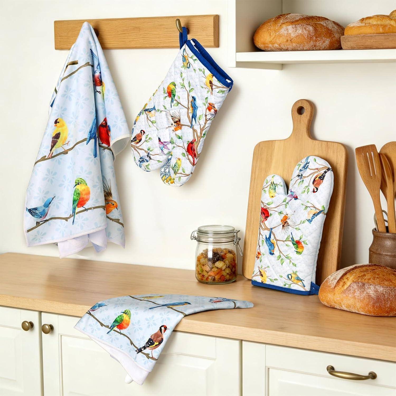 Bunte Küchentücher mit Vogelmotiven – Geschirrhandtuch, Ofenhandschuh und Topflappen – aufgehängt an Haken und auf der Arbeitsfläche neben Brot und Küchenutensilien ausgestellt.