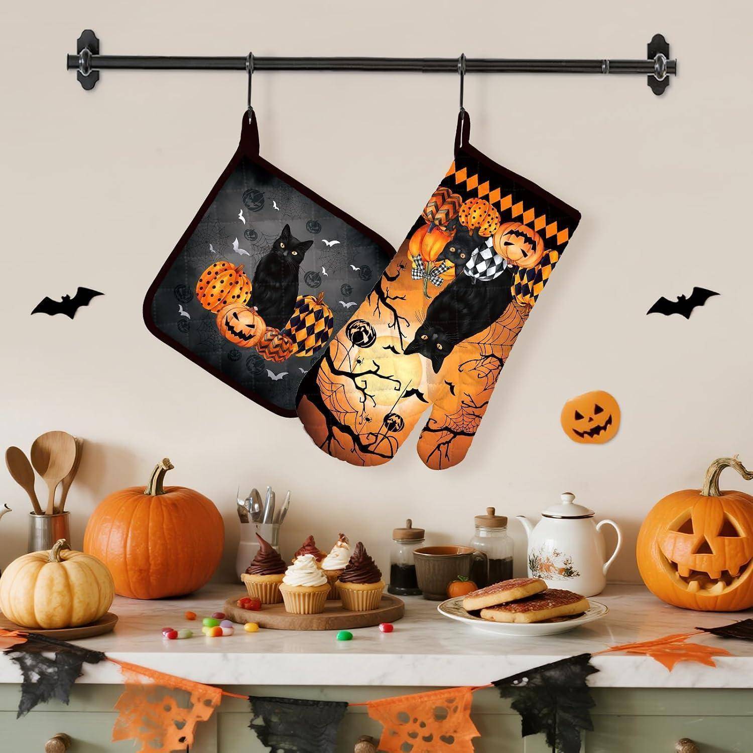 Halloween-Küchendisplay mit zwei hängenden Katzen-und-Kürbis-Ofenhandschuhen über einem Tisch mit Kürbissen, Cupcakes, Keksen und festlichen Dekorationen.