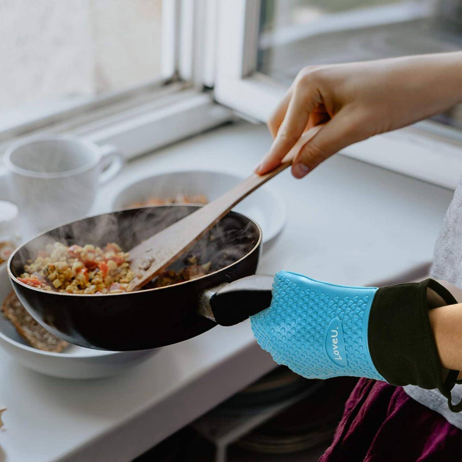 Eine Hand in einem blauen hitzebeständigen Ofenhandschuh rührt dampfendes Essen in einer Pfanne auf einer Küchentheke neben einem Fenster um.