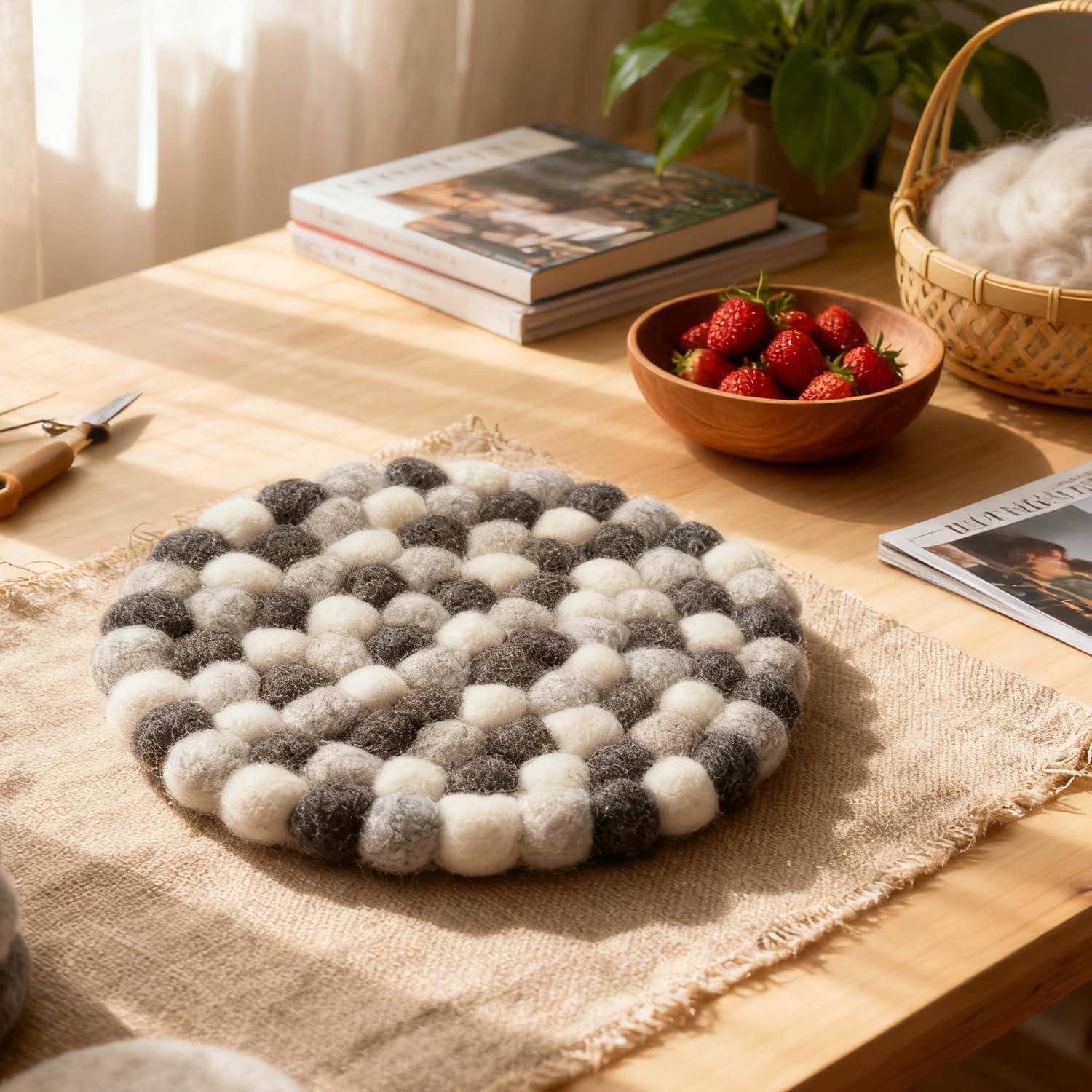 Runder grau-weißer Filzball-Untersetzer oder Matte auf einem Jute-Tuch auf einem Holztisch, mit Büchern, Erdbeeren und einem Korb in der Nähe.