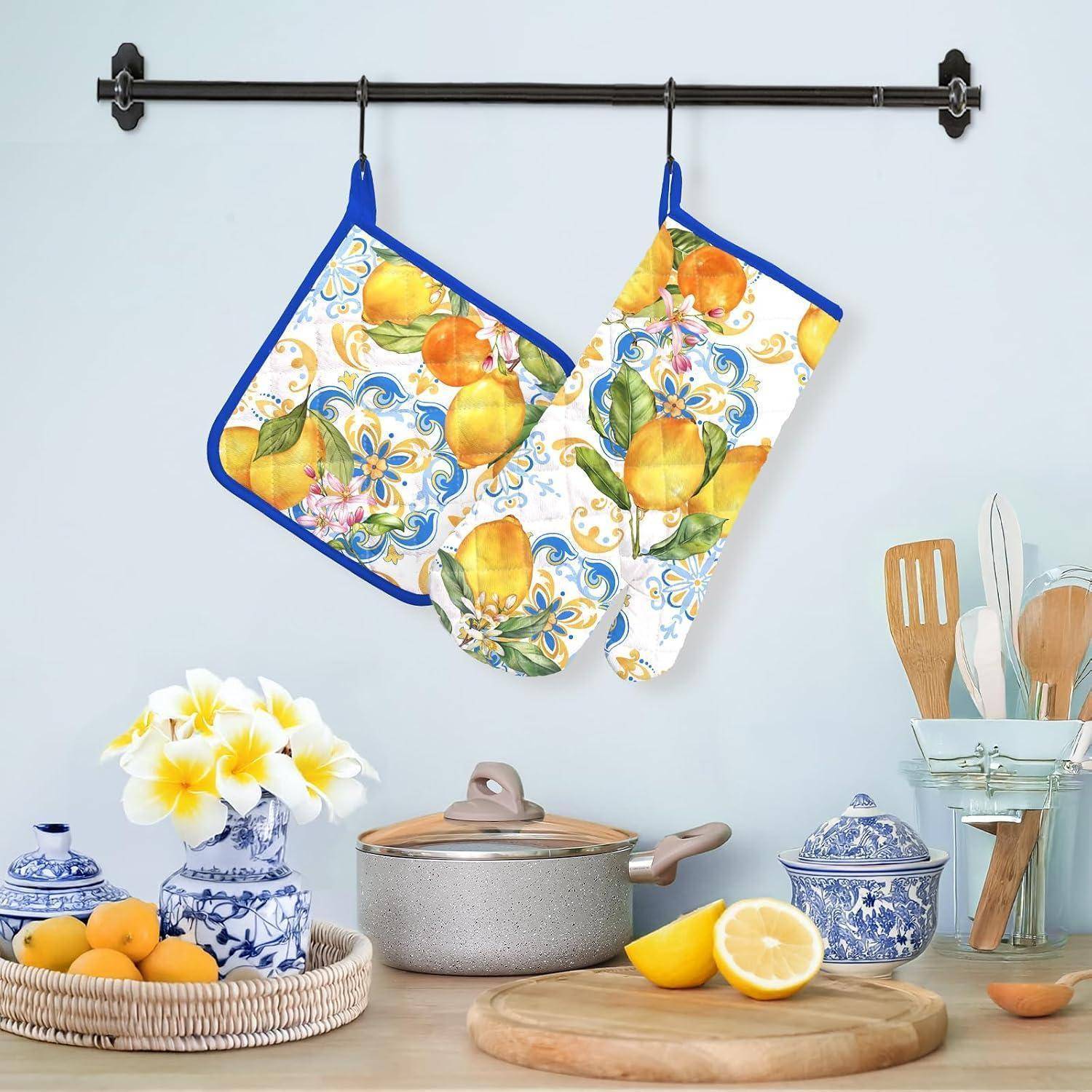 Zwei zitronenmusterierte Ofenhandschuhe hängen an einer Wandleiste über einer Küchentheke mit Kochgeschirr, Zitronen, Blumen und Utensilien.