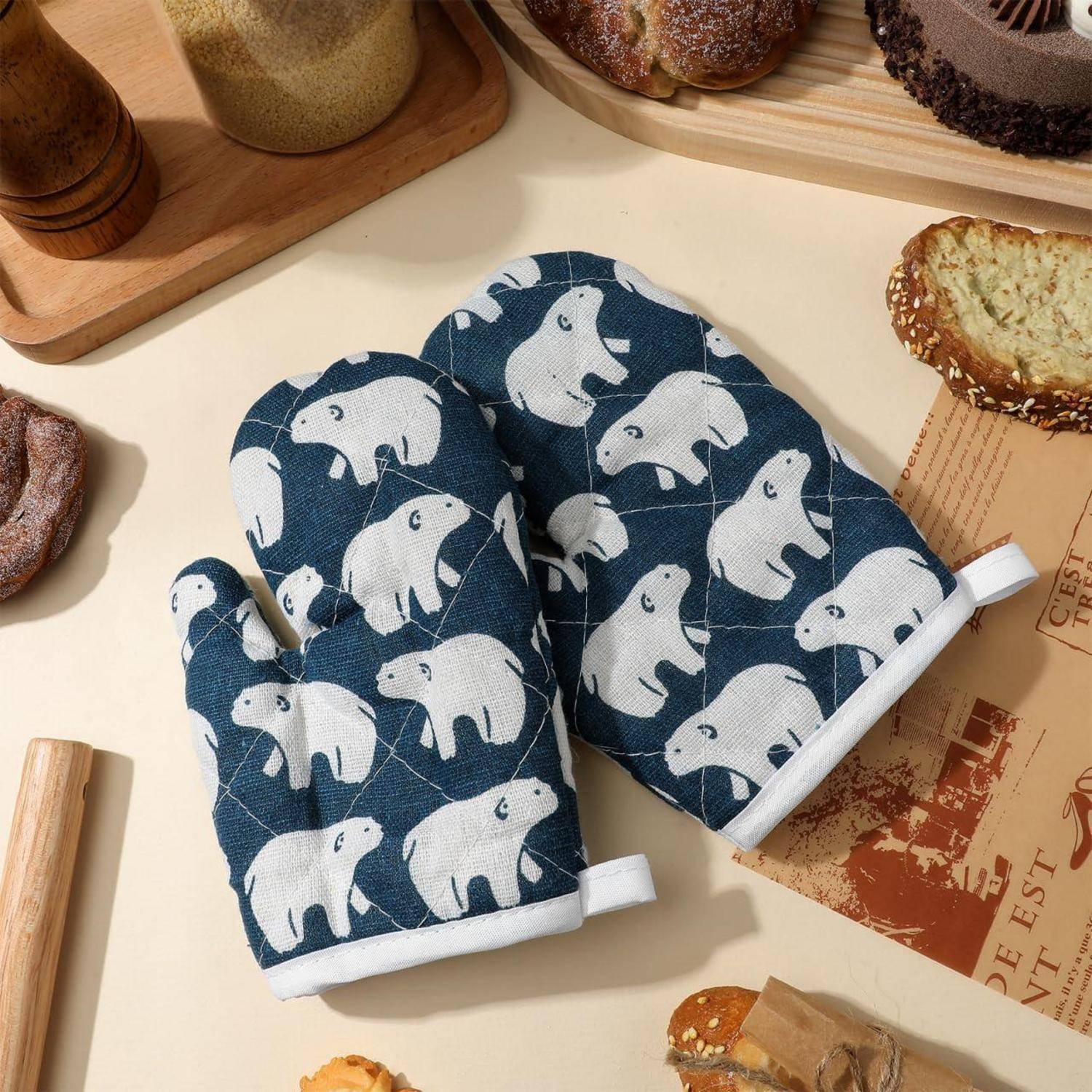 Paar marineblaue Ofenhandschuhe mit weißem Bärenmuster auf einer Tischplatte, umgeben von Brot und Backwaren.