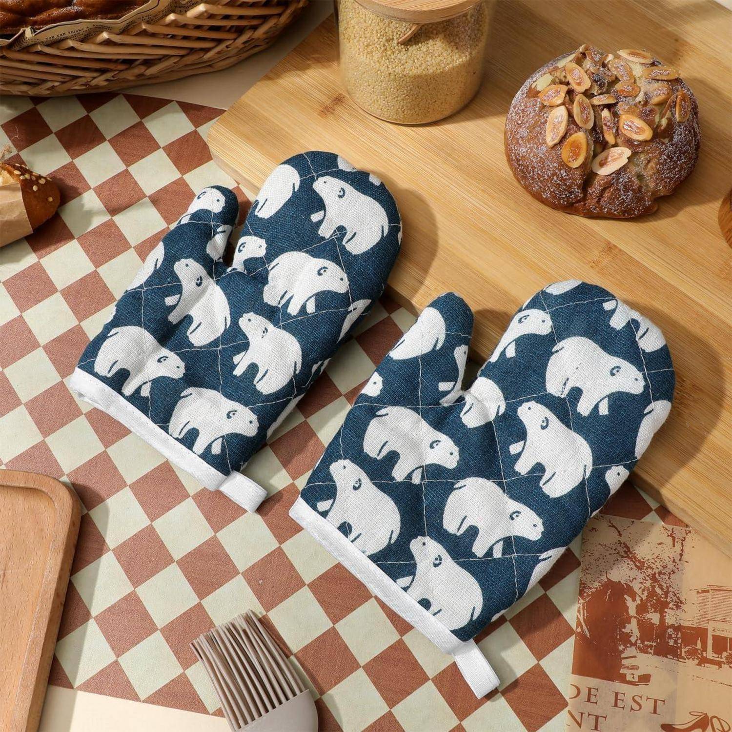 Paar blaue gesteppte Ofenhandschuhe mit weißem Eisbär-Motiv auf einem Küchentisch neben Brot und einem Glas Getreide.