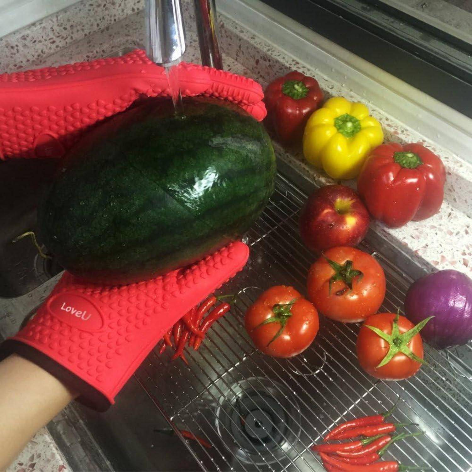 Hand hält eine dunkelgrüne Wassermelone unter fließendes Wasser in einer Küchenspüle, mit roten und gelben Paprika, Tomaten, Chilis und einer Zwiebel in der Nähe.