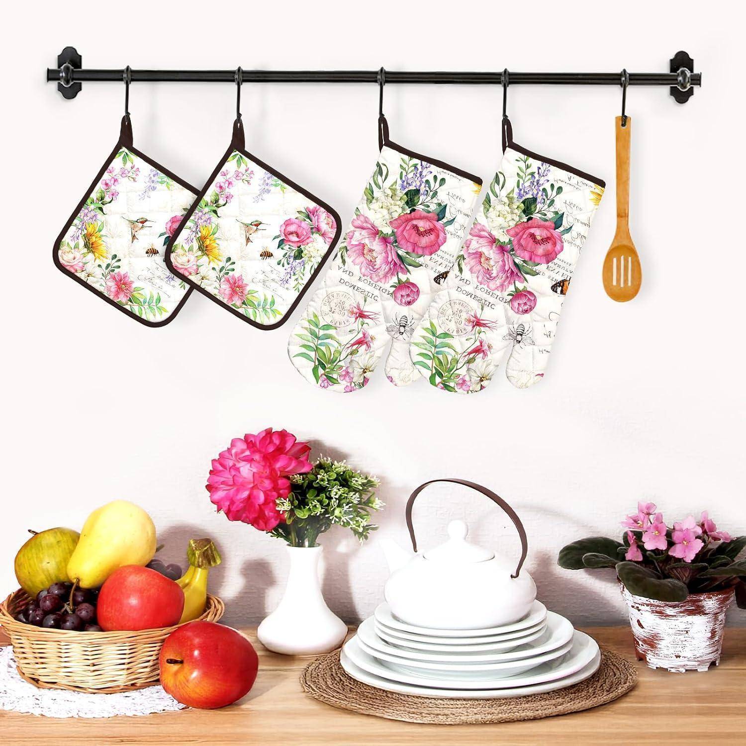 Florale Küchenzusammensetzung an einem Ständer hängend: zwei Topflappen, zwei Ofenhandschuhe und ein Holzspatel über einem Tisch mit Obst, Blumen und gestapeltem Geschirr.