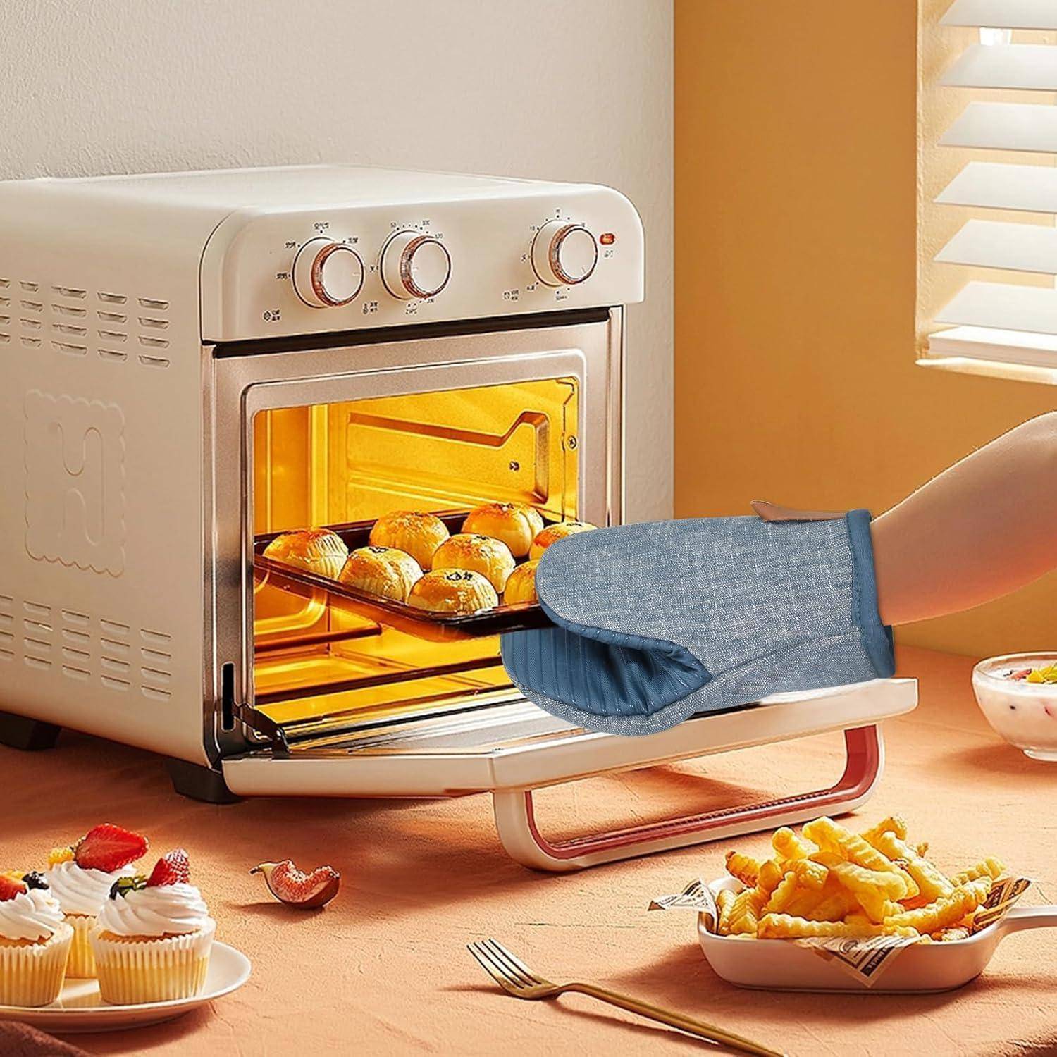 Eine Person benutzt einen Ofenhandschuh, um ein Tablett mit gebackenen Brötchen aus einem Toaster-Ofen auf der Arbeitsfläche zu ziehen, während Cupcakes und Pommes frites in der Nähe auf dem Tisch stehen.