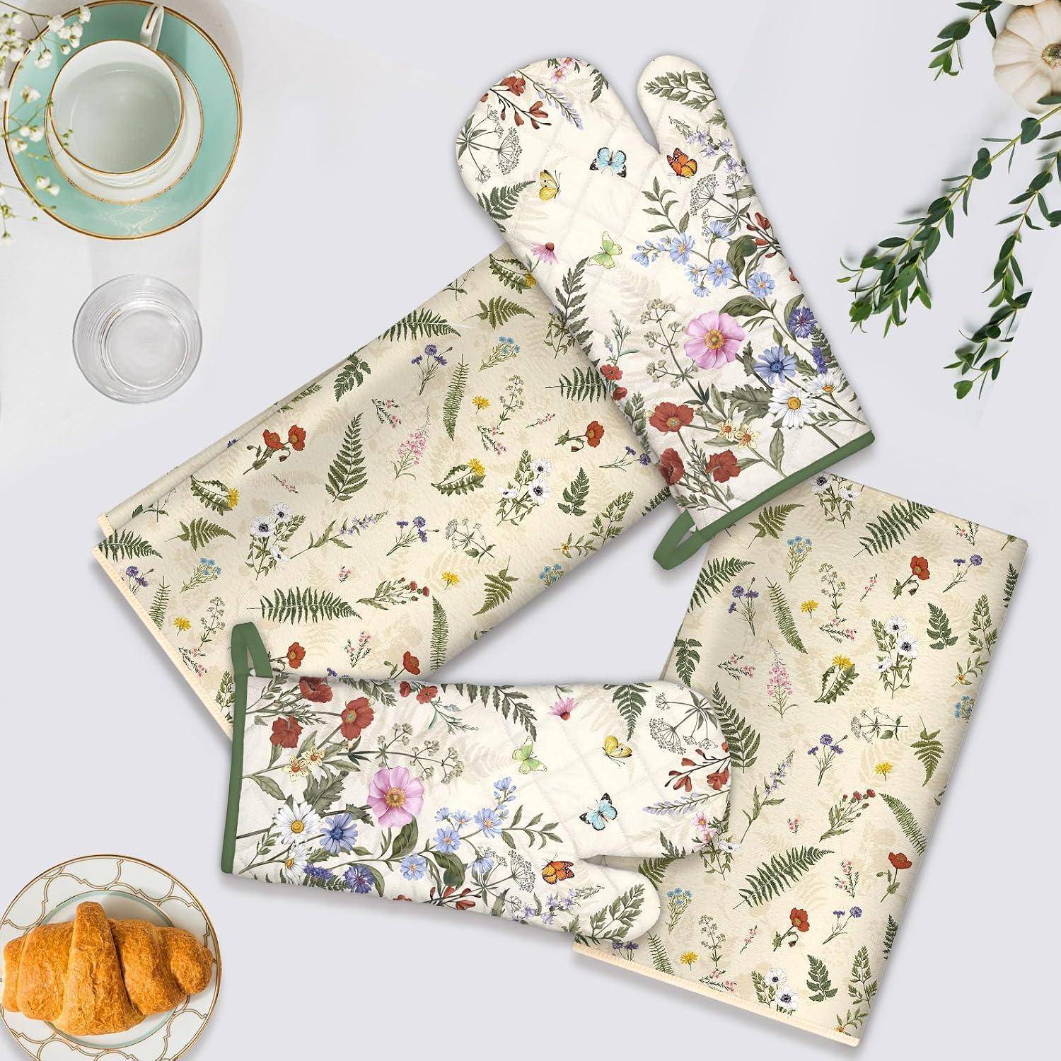 Vier mit Blumenmuster versehene Ofenhandschuhe und Topflappen auf einer hellen Oberfläche angeordnet, mit Teetassen und Croissants an den Rändern.