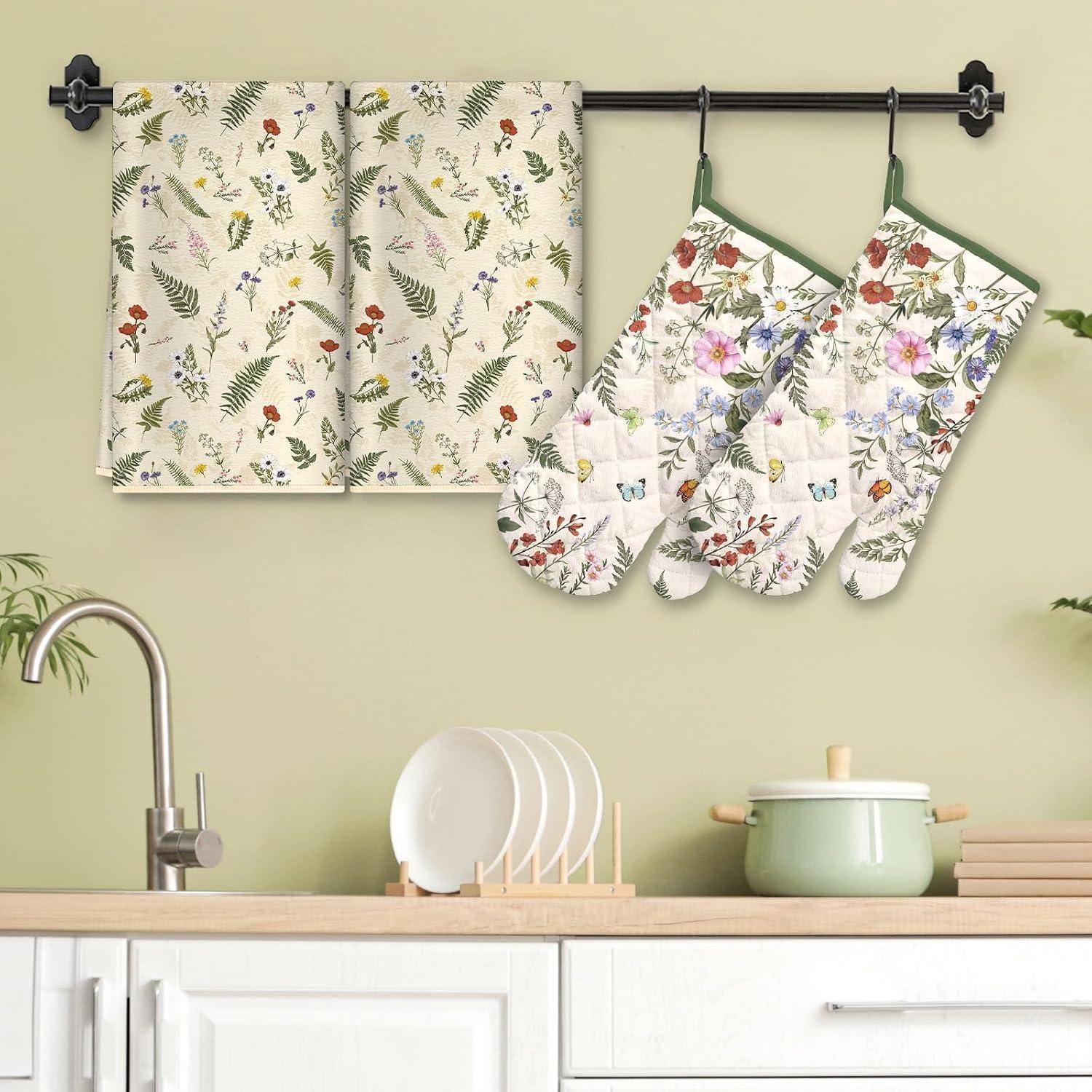 Küchentücher mit Blumenmuster und Ofenhandschuhe hängen an einer schwarzen Wandstange über einer Arbeitsplatte mit Geschirr und einem grünen Topf.