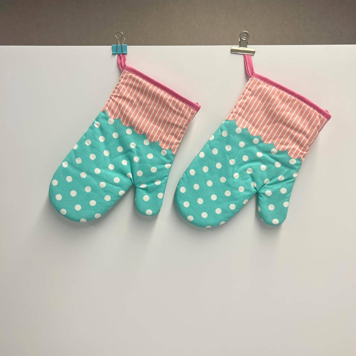 Zwei türkisfarbene Ofenhandschuhe mit weißen Pünktchen und rosa gestreiften Bündchen hängen nebeneinander an einer weißen Wand mit Klammern.