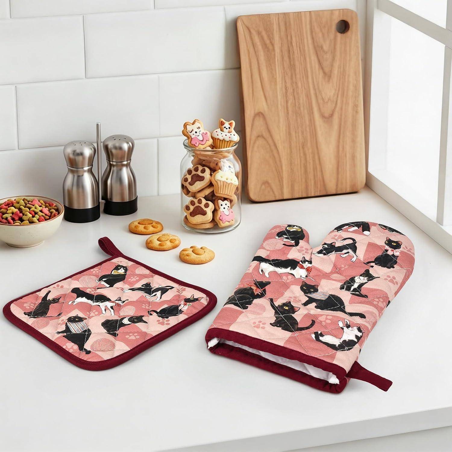 Rosa Ofenhandschuh mit Katzenmuster und passender Topflappen auf einer Küchenarbeitsplatte neben Keksen, einer Snackschüssel, Gewürzstreuern und Schneidebrettern.