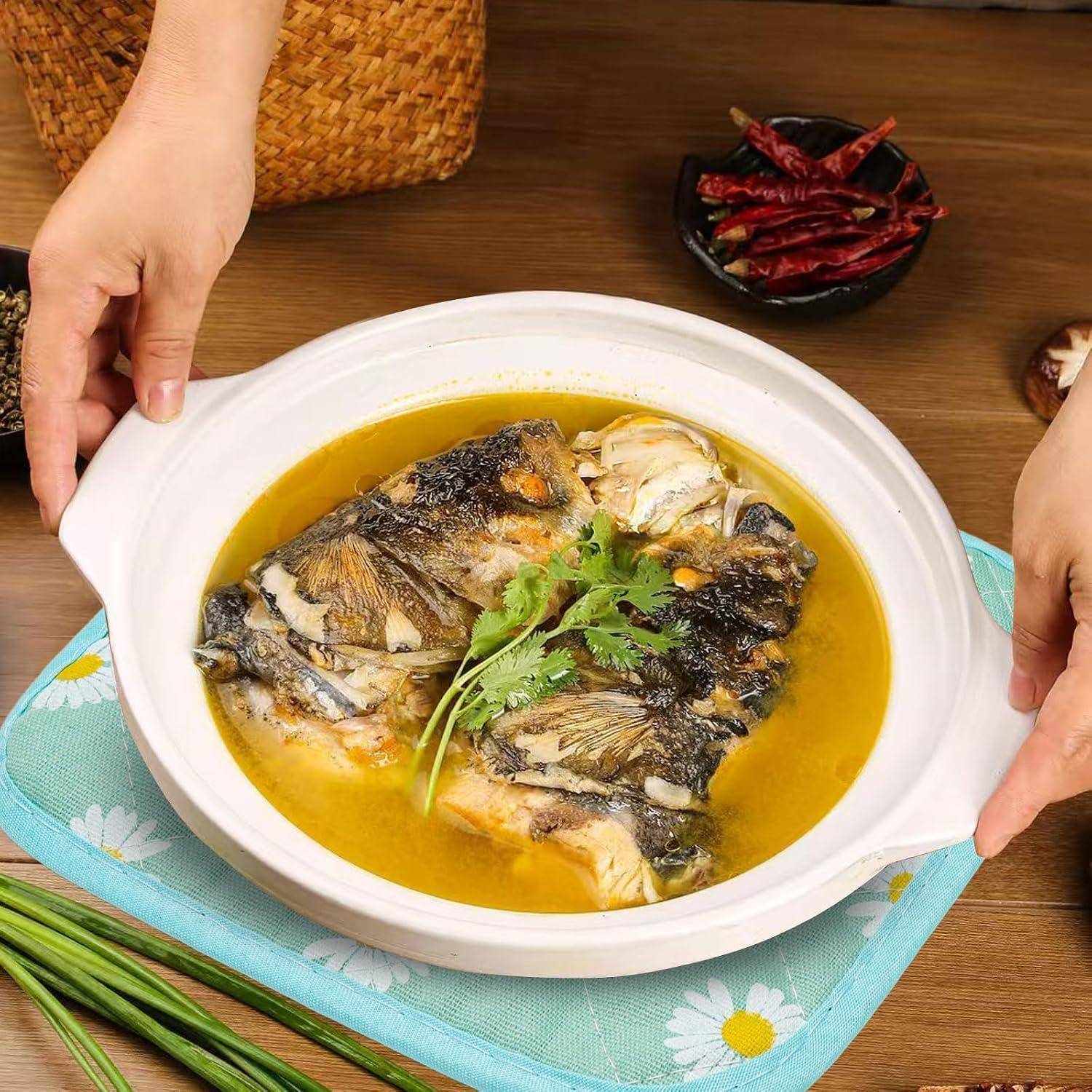 Hände platzieren eine weiße Schüssel mit Fischsuppe und Koriander auf einem Tisch, mit getrockneten Chilischoten und Frühlingszwiebeln in der Nähe.