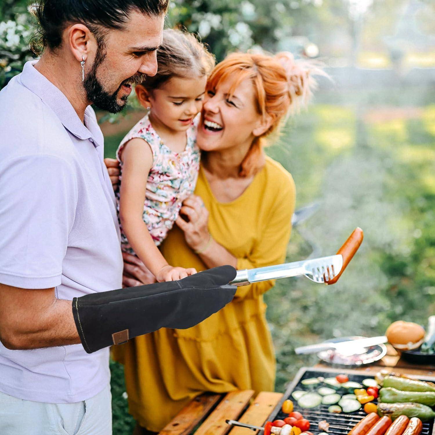 Familiengrillen im Freien: Ein Mann in einem Schutzhandschuh hält eine Wurst über einem Grill, während eine Frau in der Nähe ein Kind hält.