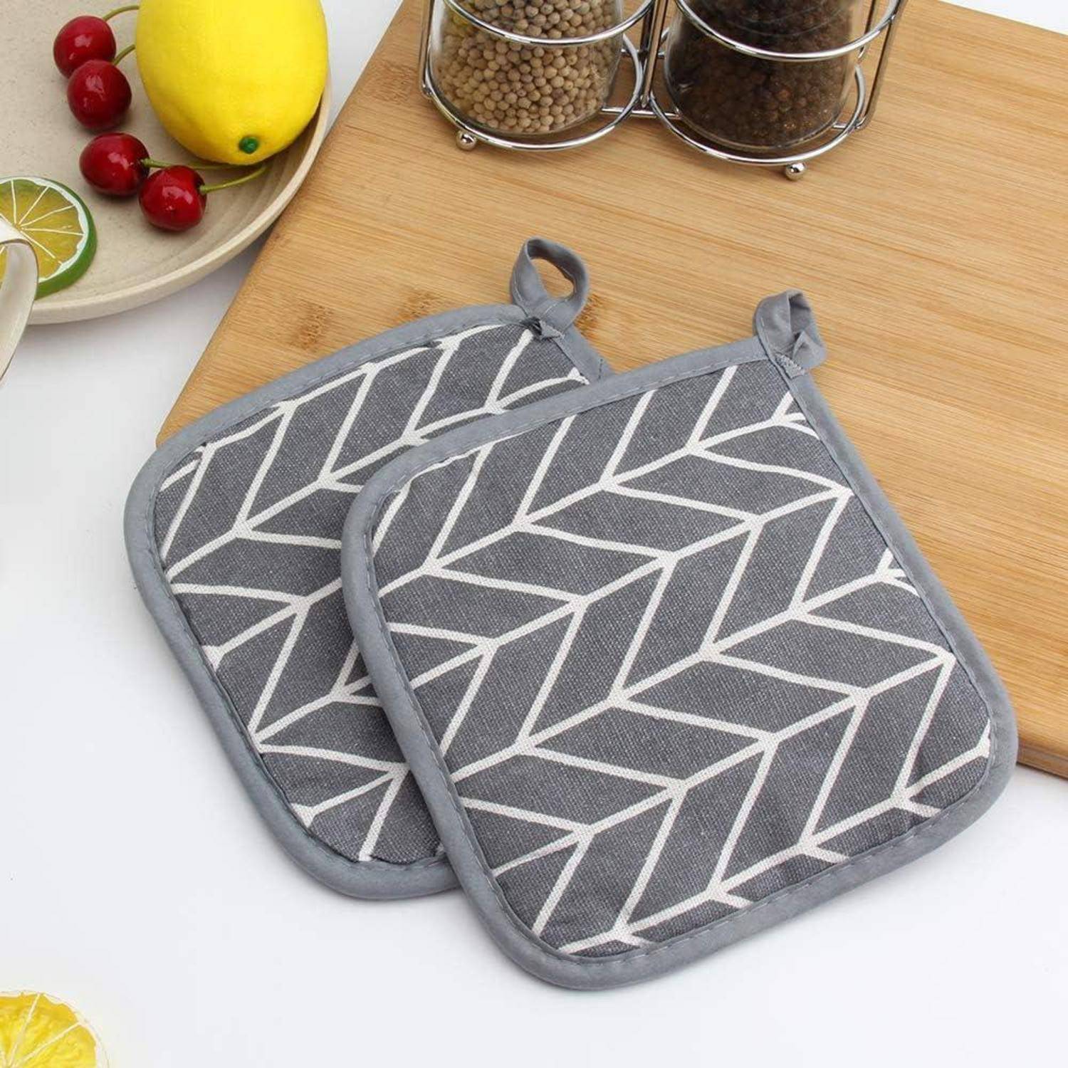 Zwei graue Topflappen mit geometrischem Muster mit Aufhängeschleifen auf einer Küchenarbeitsplatte neben Obst- und Gewürzgläsern.
