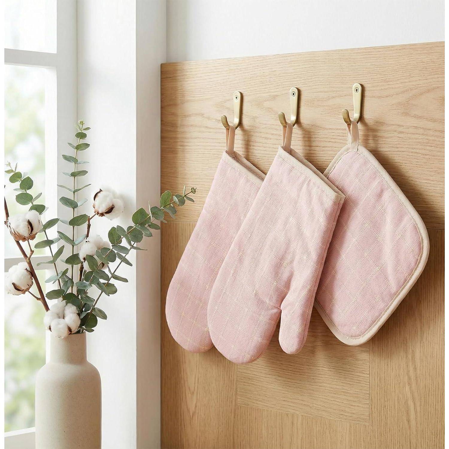 Drei rosa gesteppte Ofenhandschuhe hängen an Haken an einem Holzbrett neben einem Fenster mit einer Vase aus Baumwollstielen.