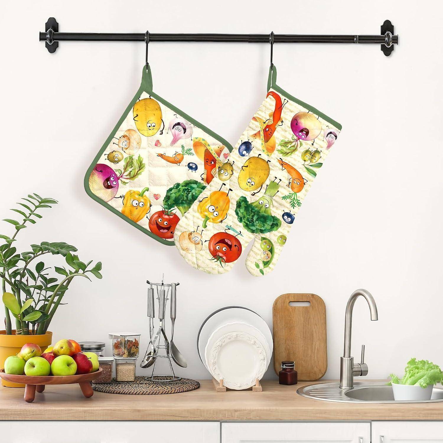 Zwei bunte gemüsemusterbedruckte Topflappen hängen an einem schwarzen Wandregal über einem Küchenbecken und einer Arbeitsplatte mit Geschirr, Obst und Utensilien.