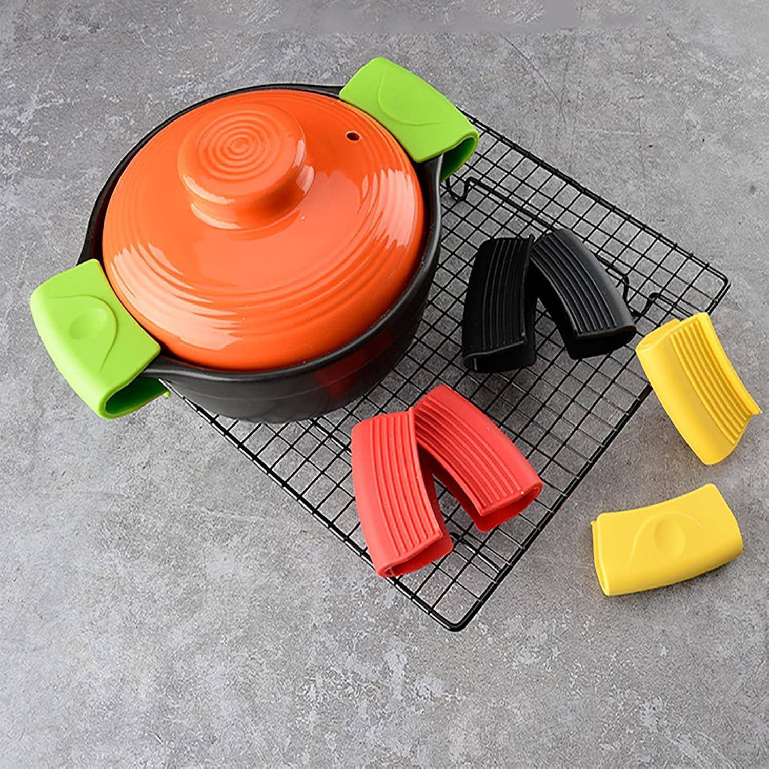 Topf mit orangefarbenem Deckel auf einem schwarzen Abkühlgitter, umgeben von farbigen Silikongriffschutzen in Grün, Schwarz, Rot und Gelb auf einer grauen Oberfläche.