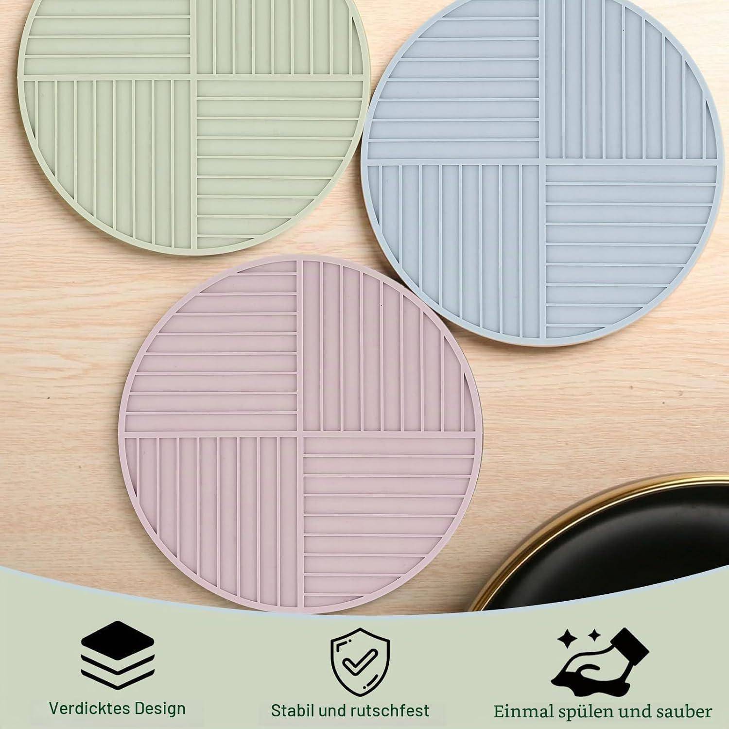 Drei runde Silikonunterlagen in Grün, Blau und Rosa auf einem Holztisch, mit Symbolen und deutschem Text darunter: „Verdicktes Design
