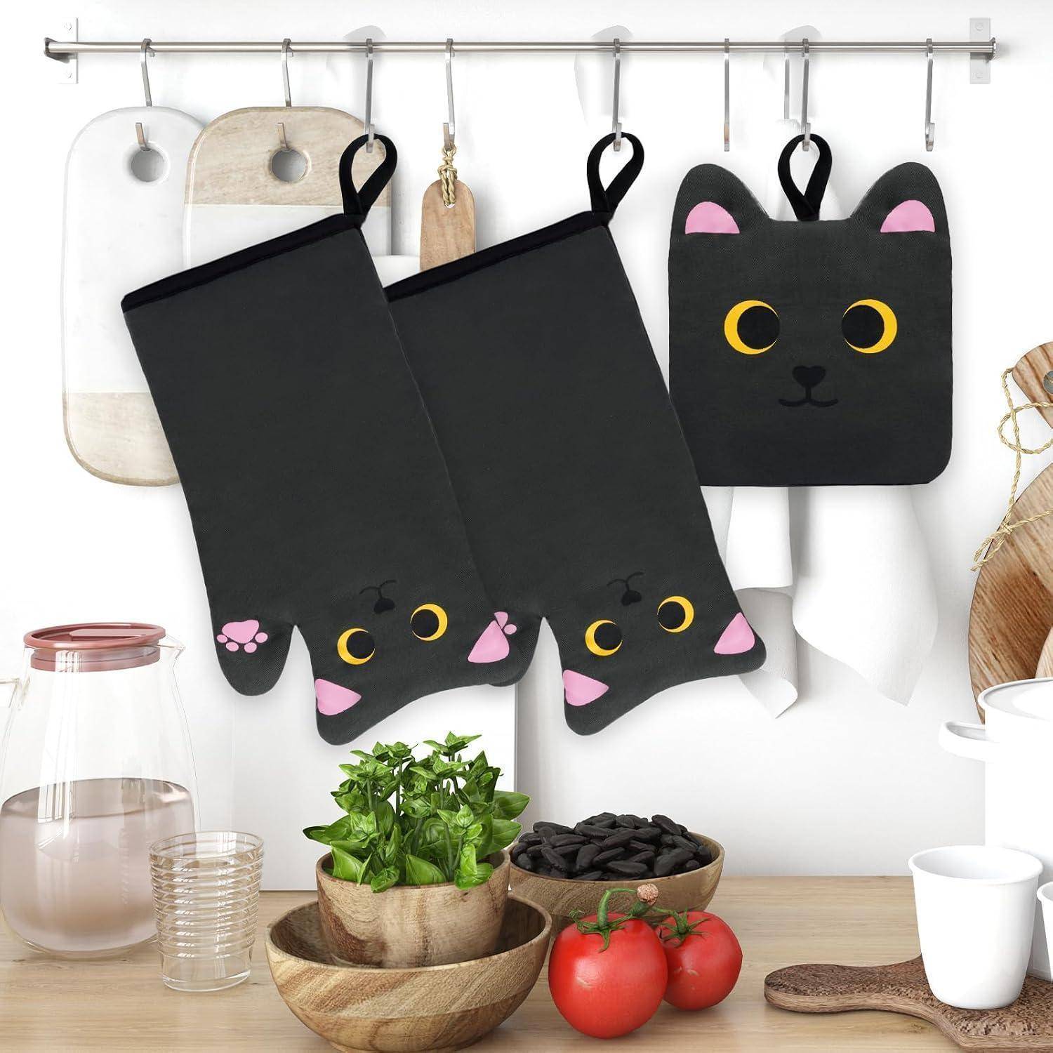 Drei schwarze katzenförmige Topflappen hängen an einem Wandregal über einer Arbeitsplatte mit Schüsseln, Tassen, einer Pflanze und Tomaten.