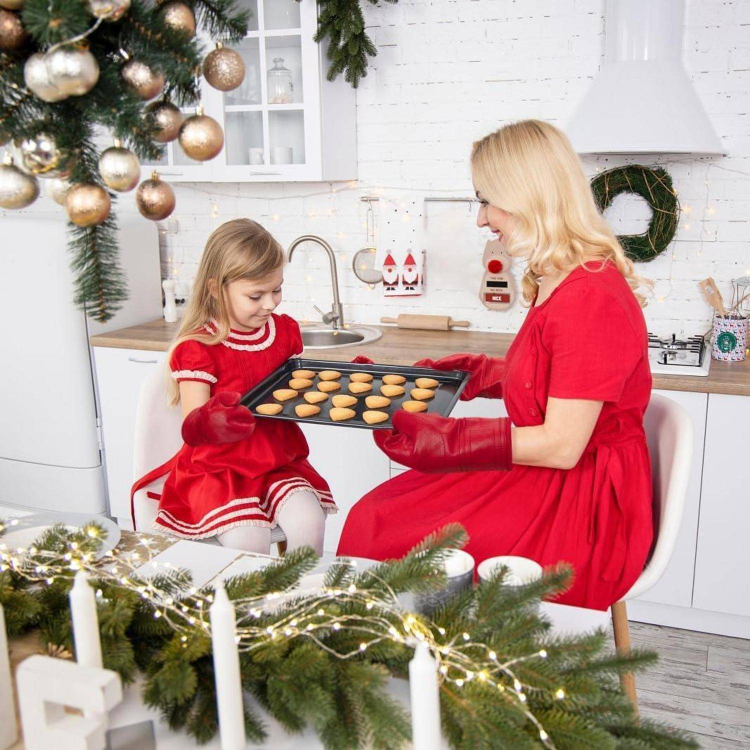 Mutter und kleine Tochter in roten Kleidern backen zusammen Kekse in einer dekorierten Weihnachtsküche und halten ein Tablett mit frisch gebackenen Keksen.