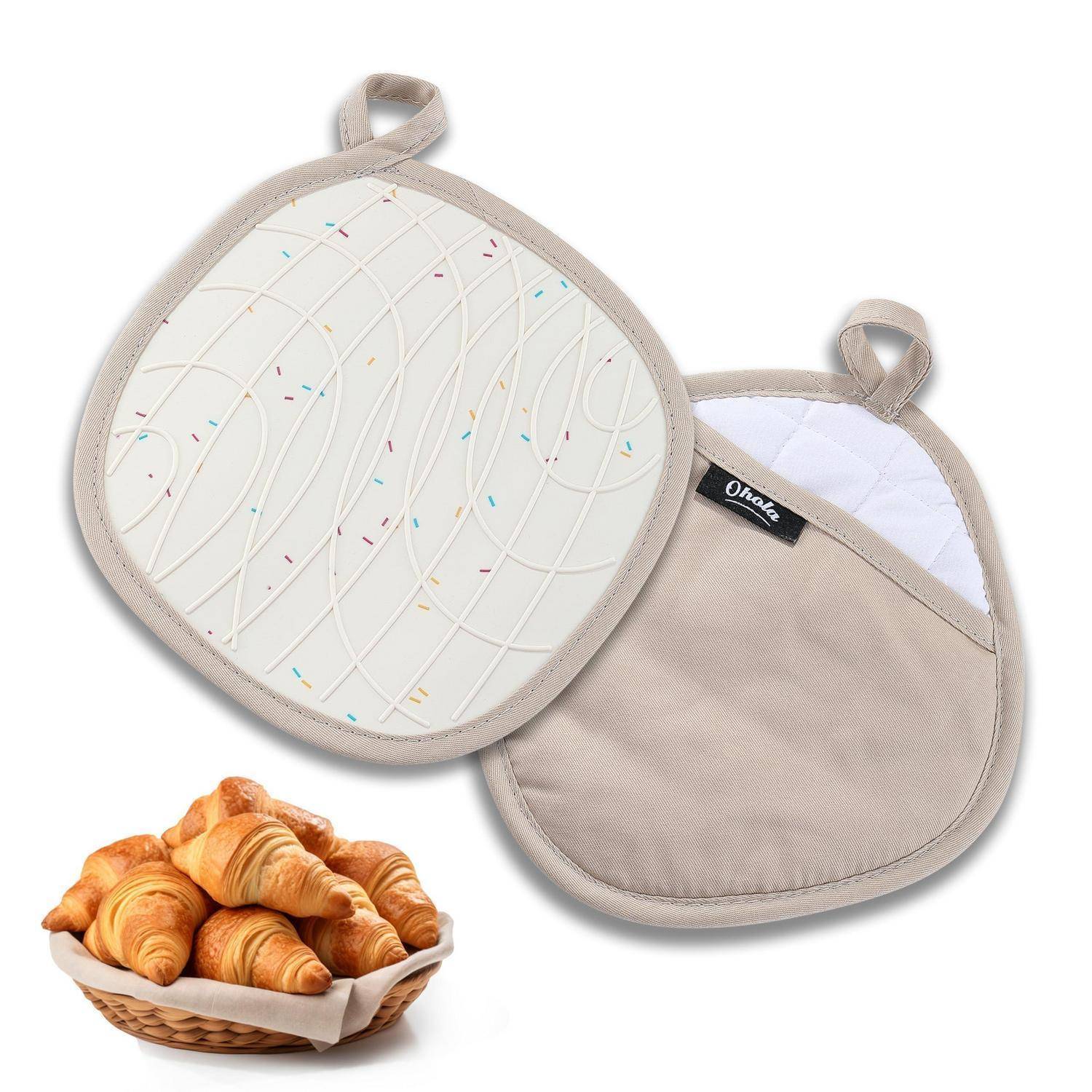 Zwei gesteppte beige Topflappen oder Ofenhandschuhe mit Aufhängeschleifen, einer mit gemusterter Vorderseite und einer schlichten Rückseite, neben einem Korb mit Croissants.