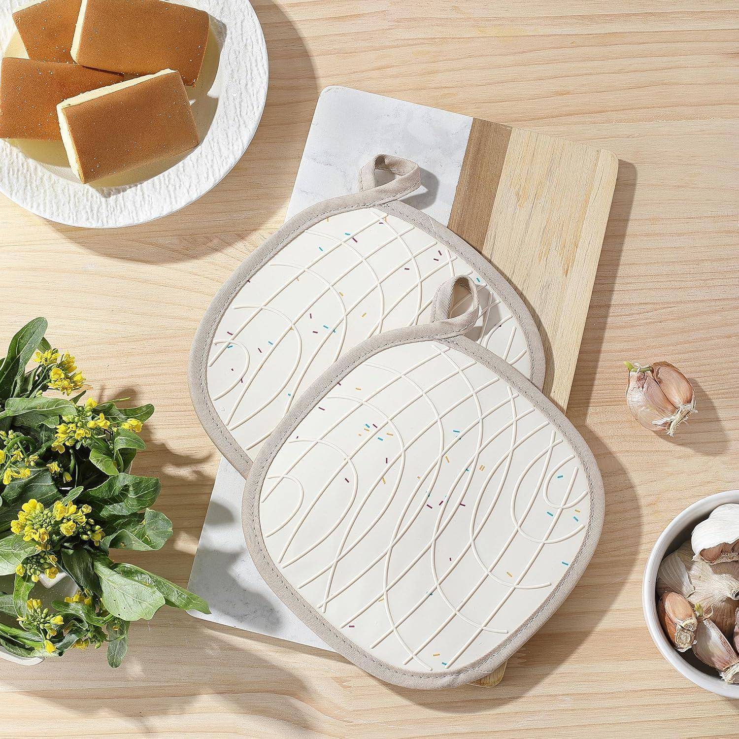 Zwei gesteppte weiße Ofenhandschuhe mit Schleifen zum Aufhängen auf einem Holzschneidebrett, umgeben von Brot auf einem Teller, Knoblauch und Topfblumen.