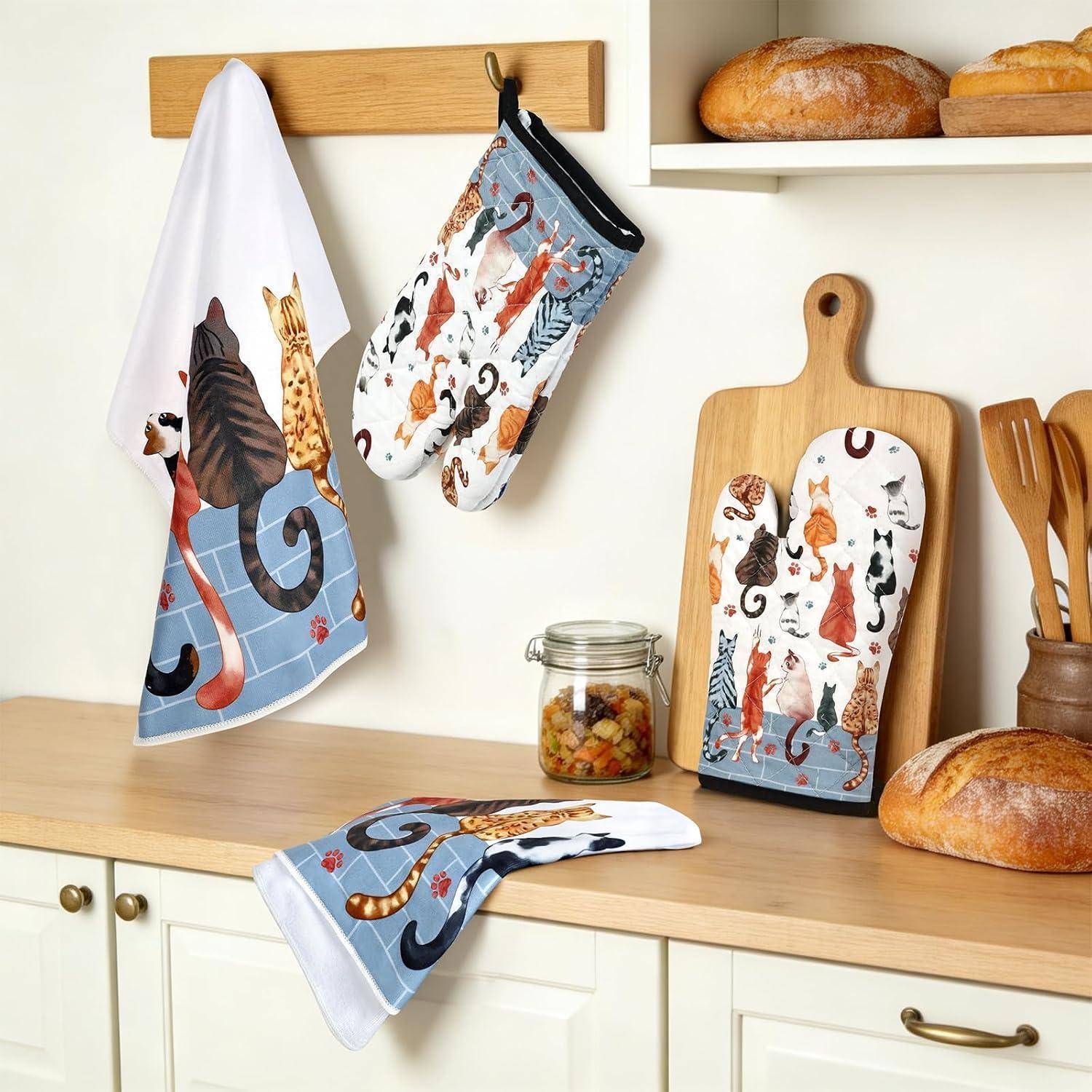 Küchenszene mit katzenmustrigen Ofenhandschuhen und passendem Handtuch, die an Haken aufgehängt sind, sowie ein Schneidebrett neben Brot und Utensilien.