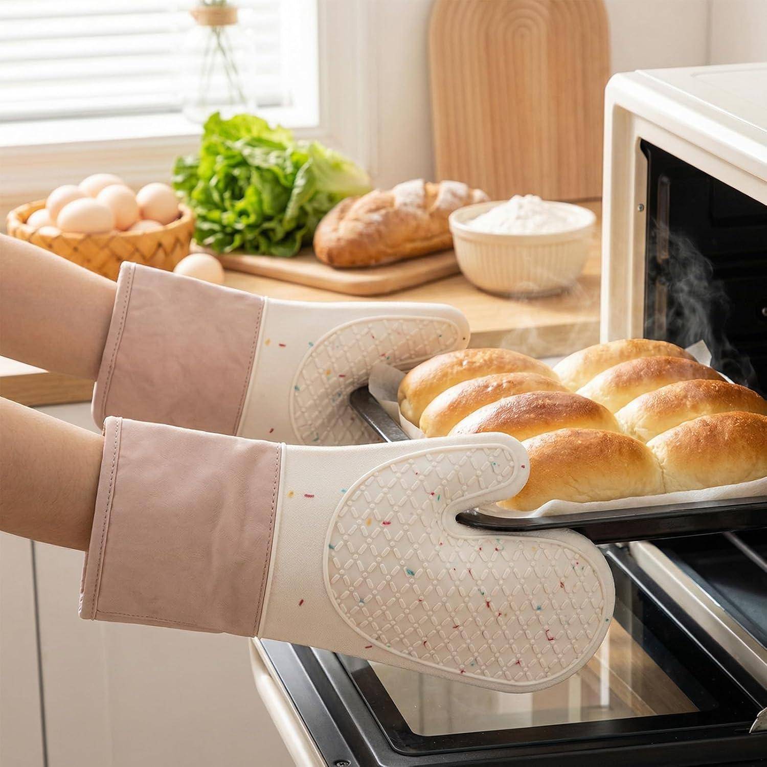 Hände in Ofenhandschuhen ziehen ein Blech mit frisch gebackenen Brötchen aus einem Ofen in einer hellen Küche, wobei Zutaten auf der Arbeitsfläche dahinter liegen.