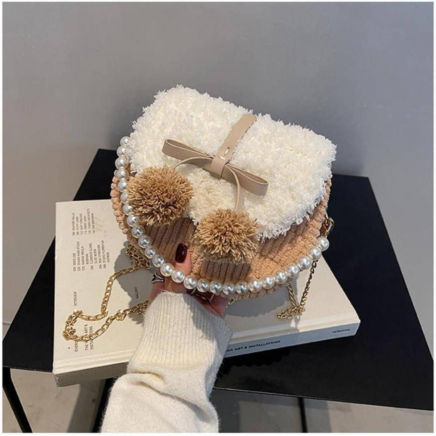 Hand hält eine kleine flauschige herzförmige beige Handtasche mit Perleneinfassung, Schleife und Pompons über einem Buch auf einem schwarzen Tisch.