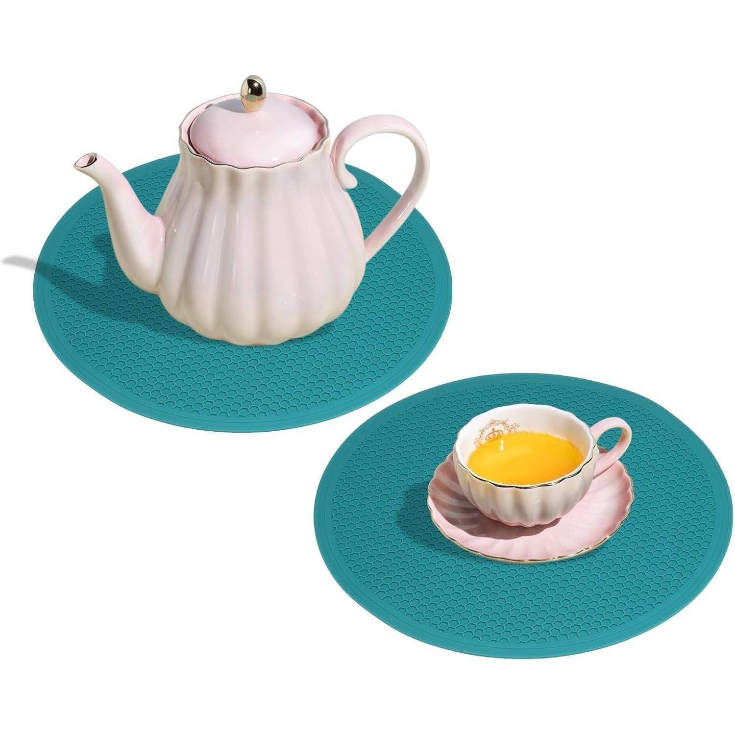 Rosa Porzellanteekanne und Teetasse auf passenden Untertassen, jeweils auf einem türkisfarbenen runden Platzset vor weißem Hintergrund platziert.