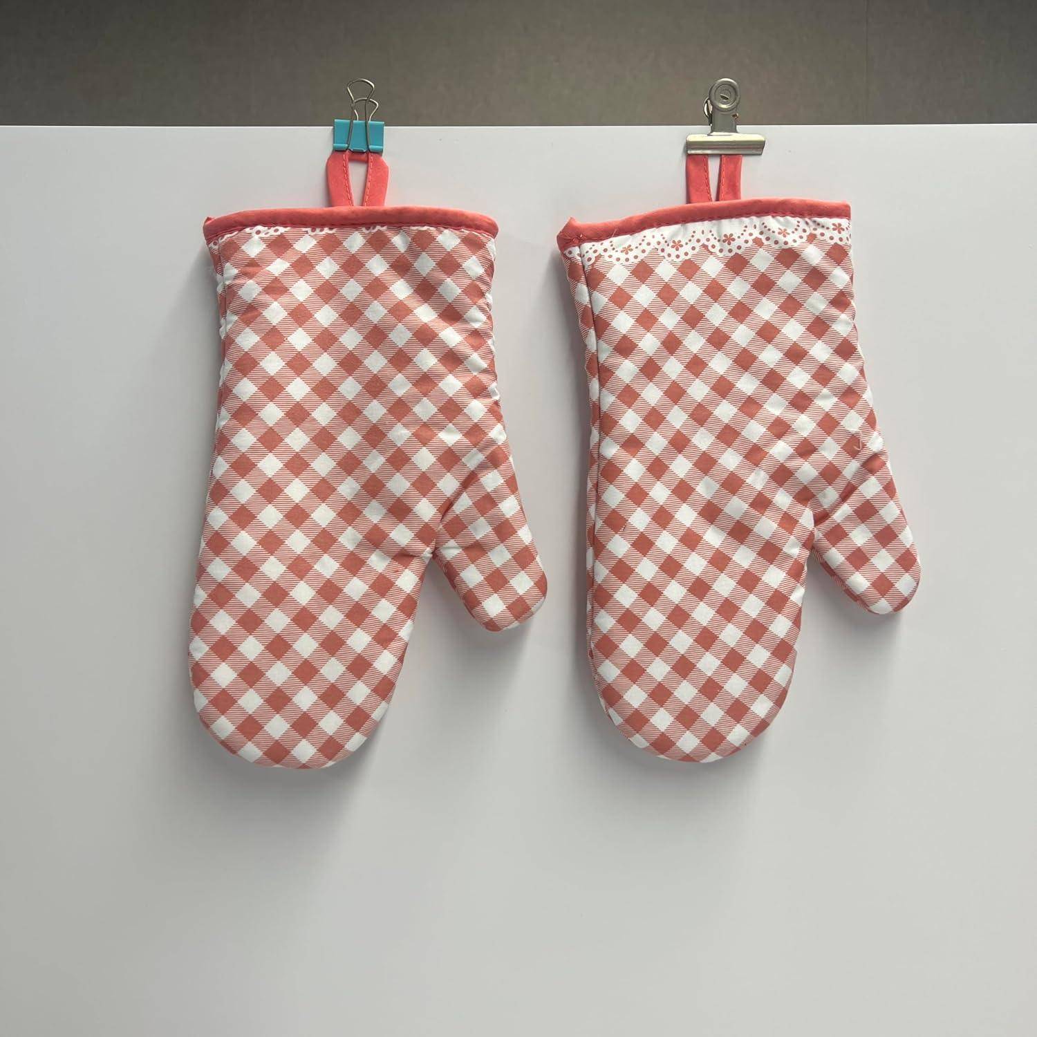 Zwei rot-weiß karierte Ofenhandschuhe hängen nebeneinander auf Clips an einer schlichten, hellen Wand.