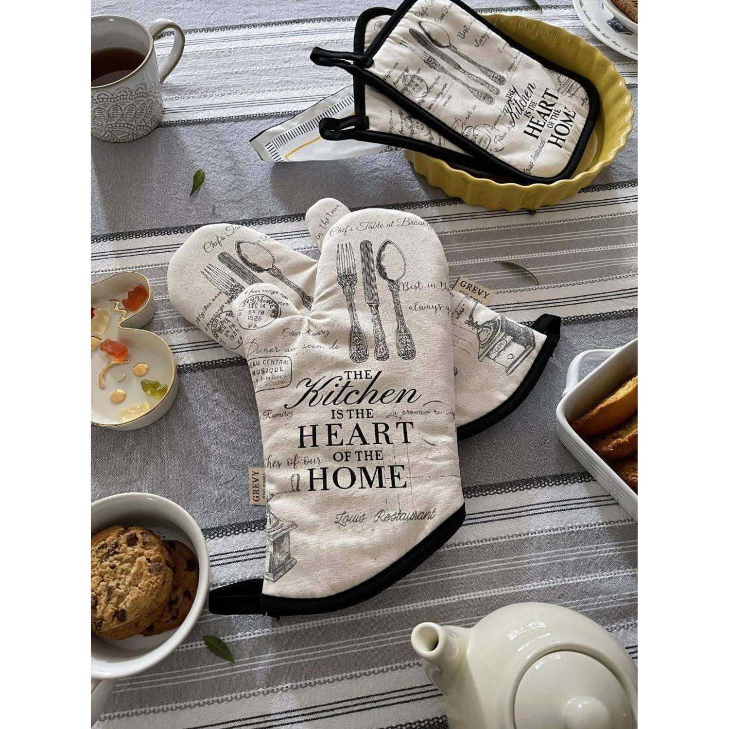 Küchenthematische Ofenhandschuhe und Topflappen auf einem Tisch, bedruckt mit „The Kitchen is the Heart of the Home