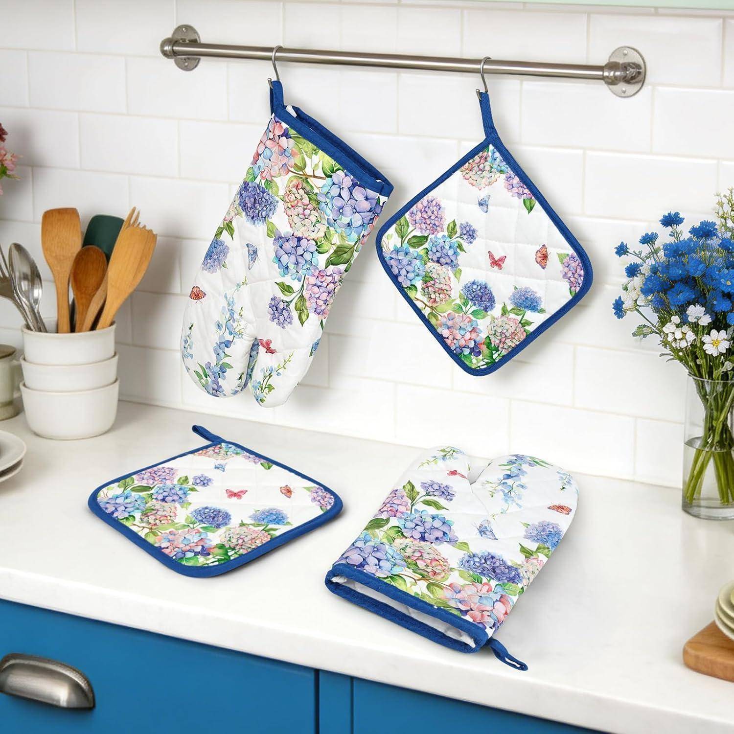Textilienset für die Küche mit Blumenmuster, bestehend aus zwei Ofenhandschuhen und zwei Topflappen, die auf einer weißen Arbeitsplatte in einer hellen Küche hängend und ausgelegt sind.