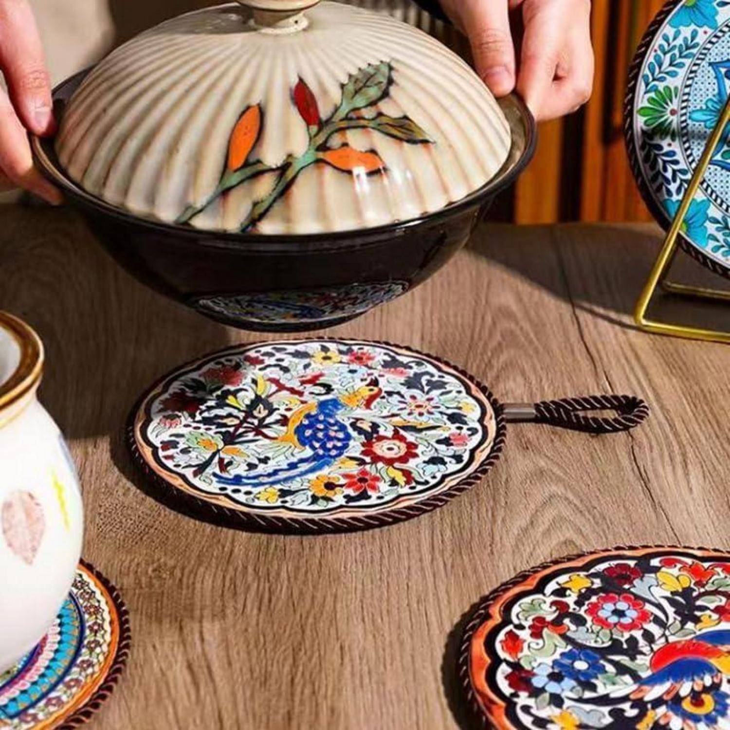 Hände heben eine gemusterte Keramik-Servierschüssel mit Deckel über einen Tisch, auf dem bunte dekorative Teller und Untersetzer ausgestellt sind.