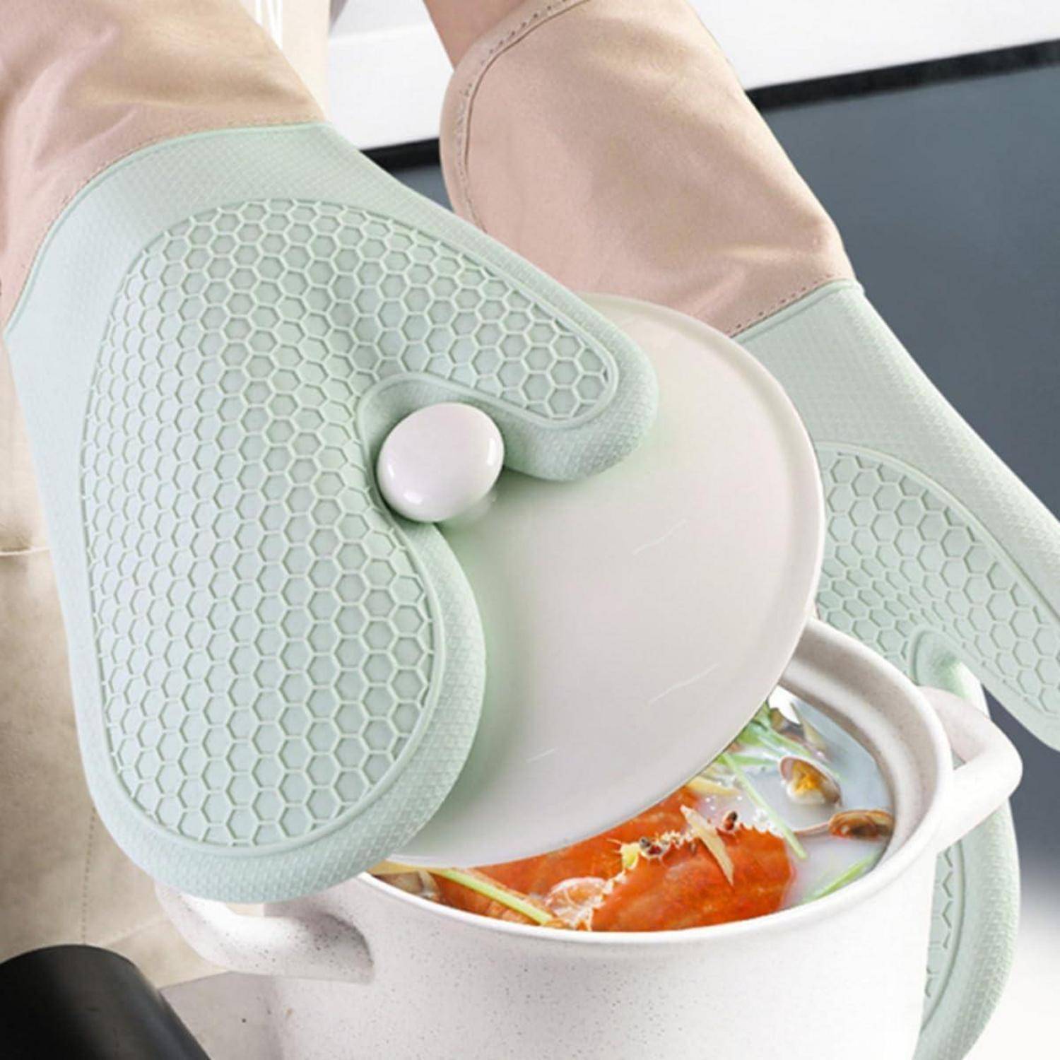 Hände mit mintgrünen hitzebeständigen Silikon-Topflappen, die den Deckel eines weißen Topfes mit dampfender Suppe heben.