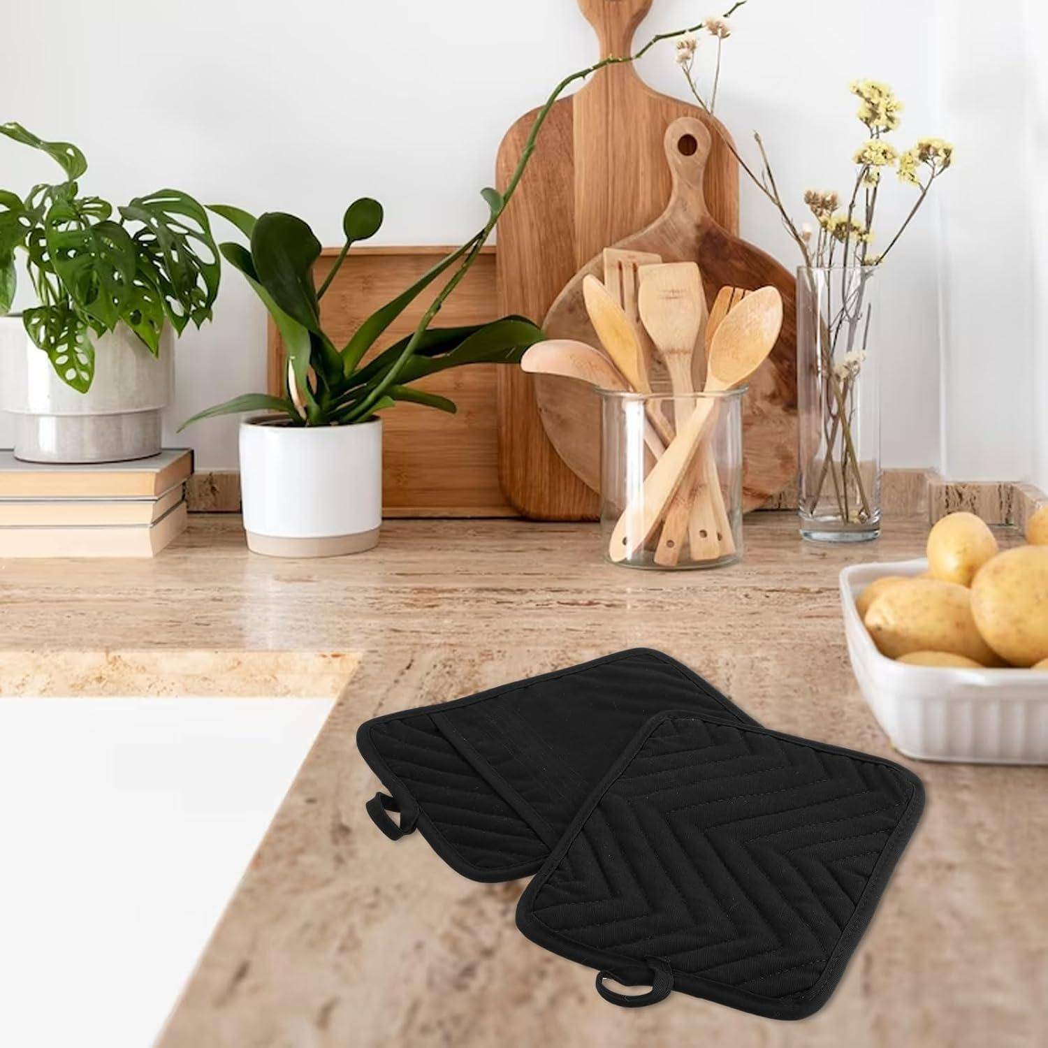 Schwarze gesteppte Topflappen auf einer Küchentheke neben Holzutensilien, Topfpflanzen und einer Schüssel mit Zitronen.