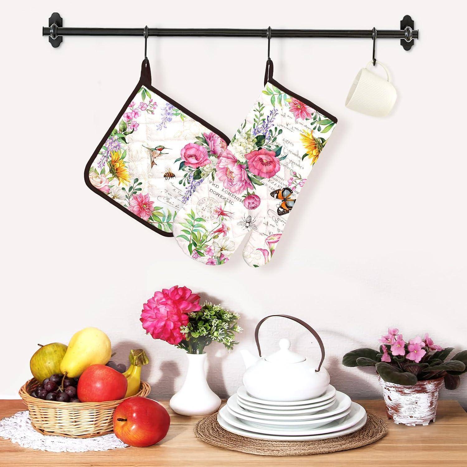 Bunte Ofenhandschuhe, Topflappen und eine Schürze hängen an einer Wand über einem Tisch, der mit Obst, Blumen, Geschirr und einer Teekanne gedeckt ist.