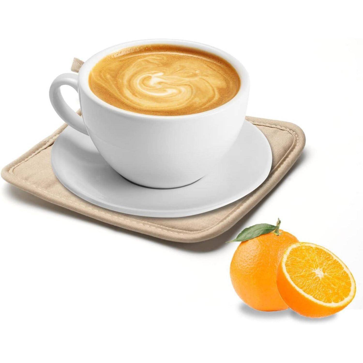 Eine Tasse Latte auf einer Untertasse und einen beigen Untersetzer neben zwei Orangen, eine davon aufgeschnitten, auf weißem Hintergrund.