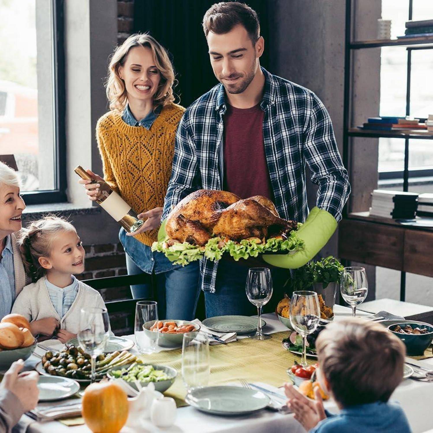 Ein Mann hält einen gebratenen Truthahn, während er eine Familie bedient, die um einen Esstisch zu einem Thanksgiving-Essen versammelt ist.