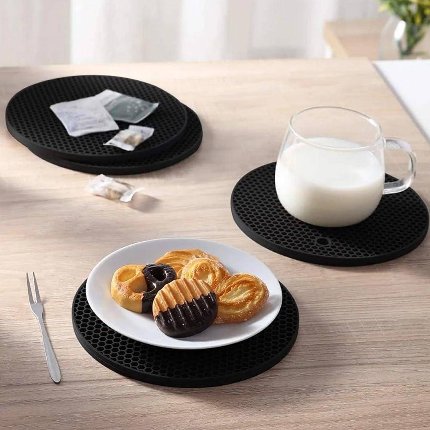 Eine Tasse Milch und ein Teller mit Gebäck auf schwarzen runden Untersetzern auf einem Holztisch, mit einer Gabel und Teebeuteln in der Nähe.