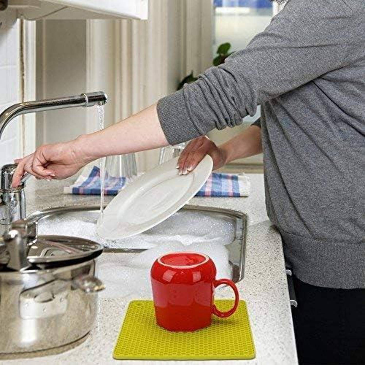 Person wäscht Geschirr in einer Küchenspüle, spült einen weißen Teller neben einer roten Tasse und einem Topf auf der Arbeitsplatte ab.